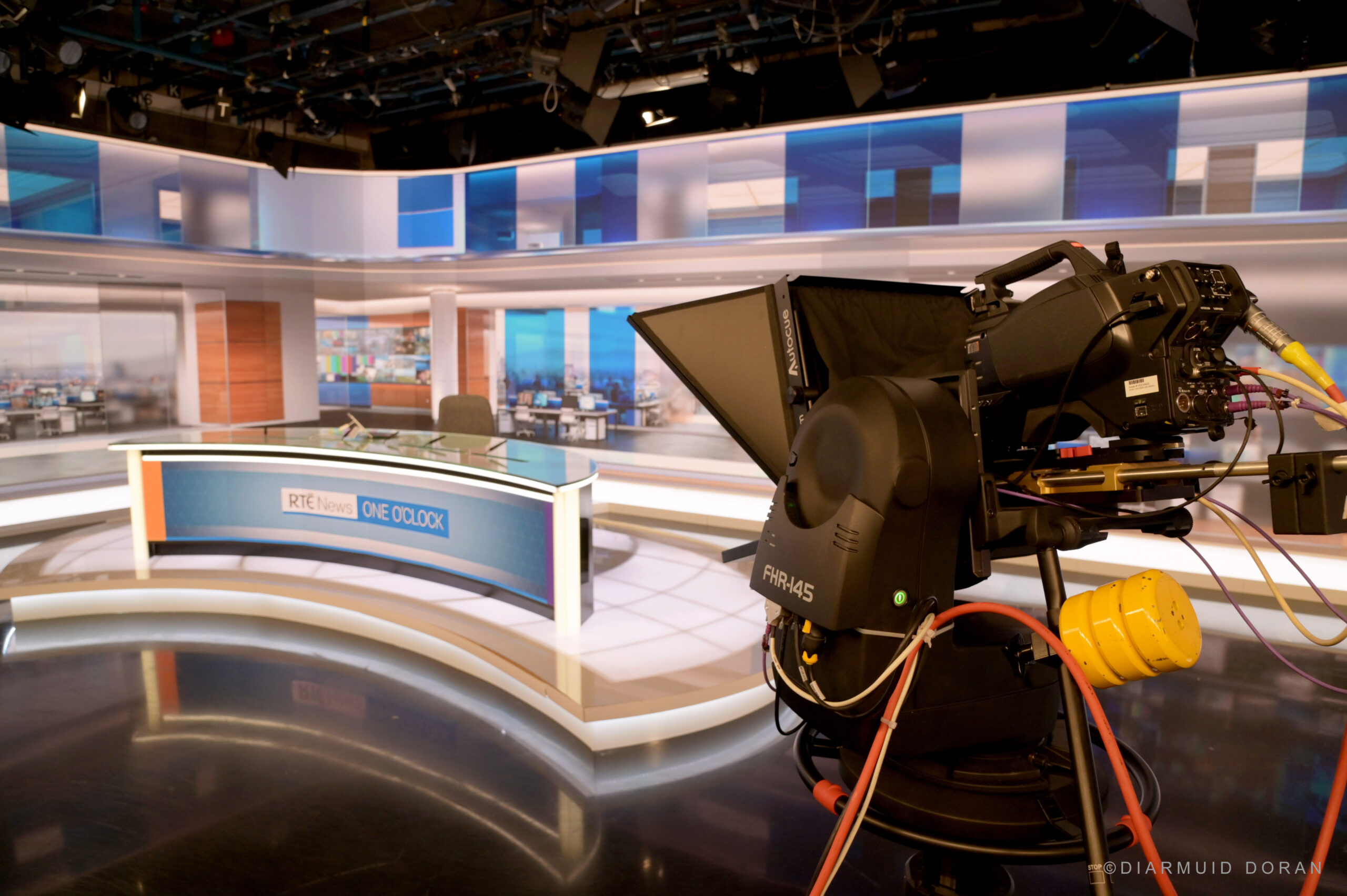 A camera in the foreground while a TV news studio is in the background.