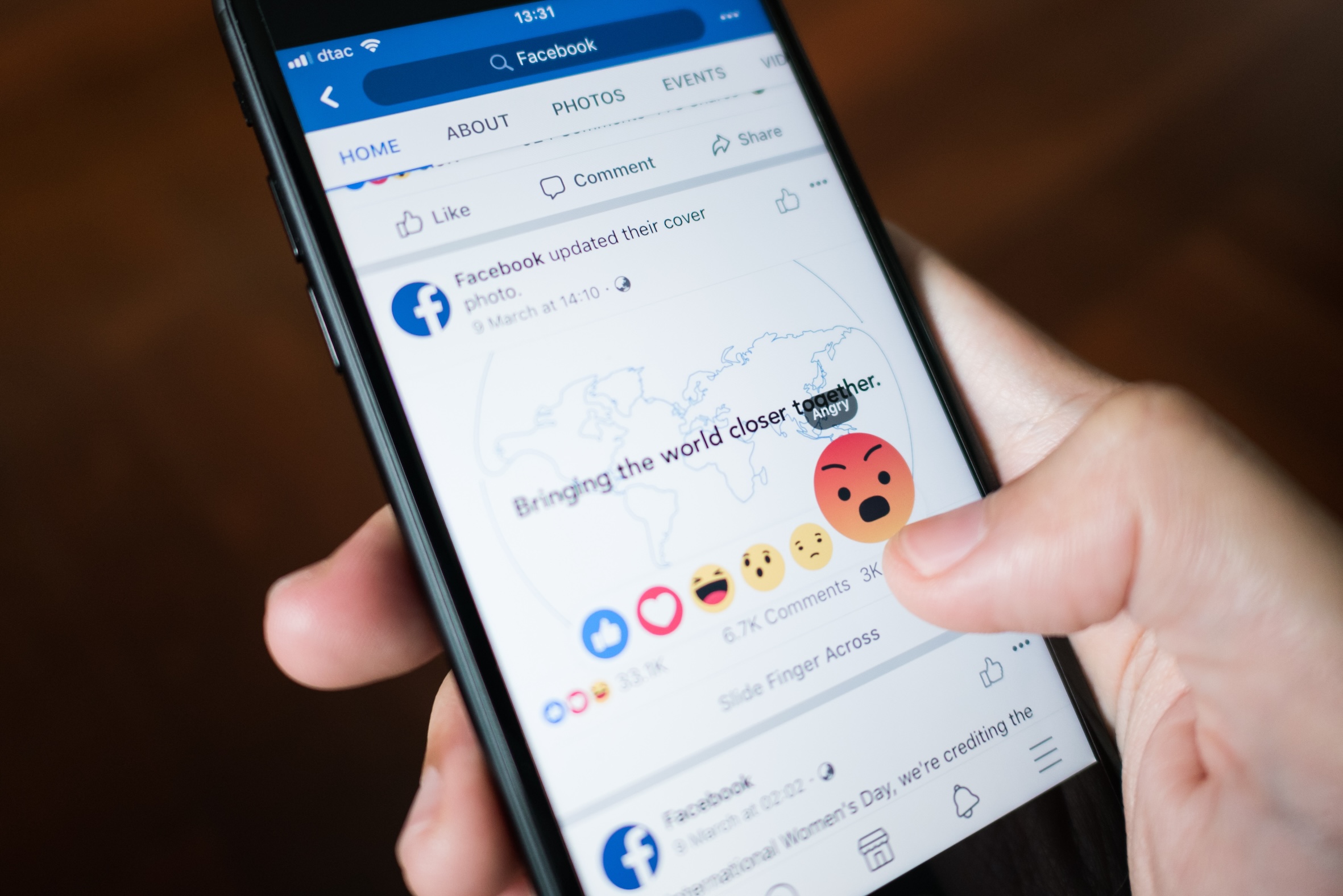 A person holds a phone, with their finger holding the angry face emoji response on Facebook