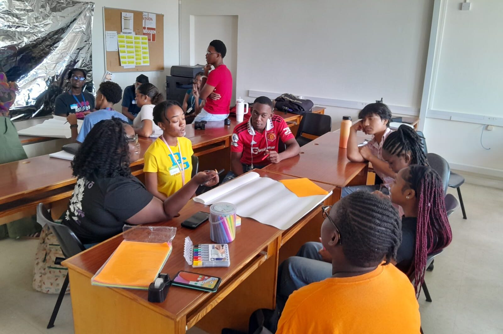 Two groups of children sit around tables with pieces of paper and pens.